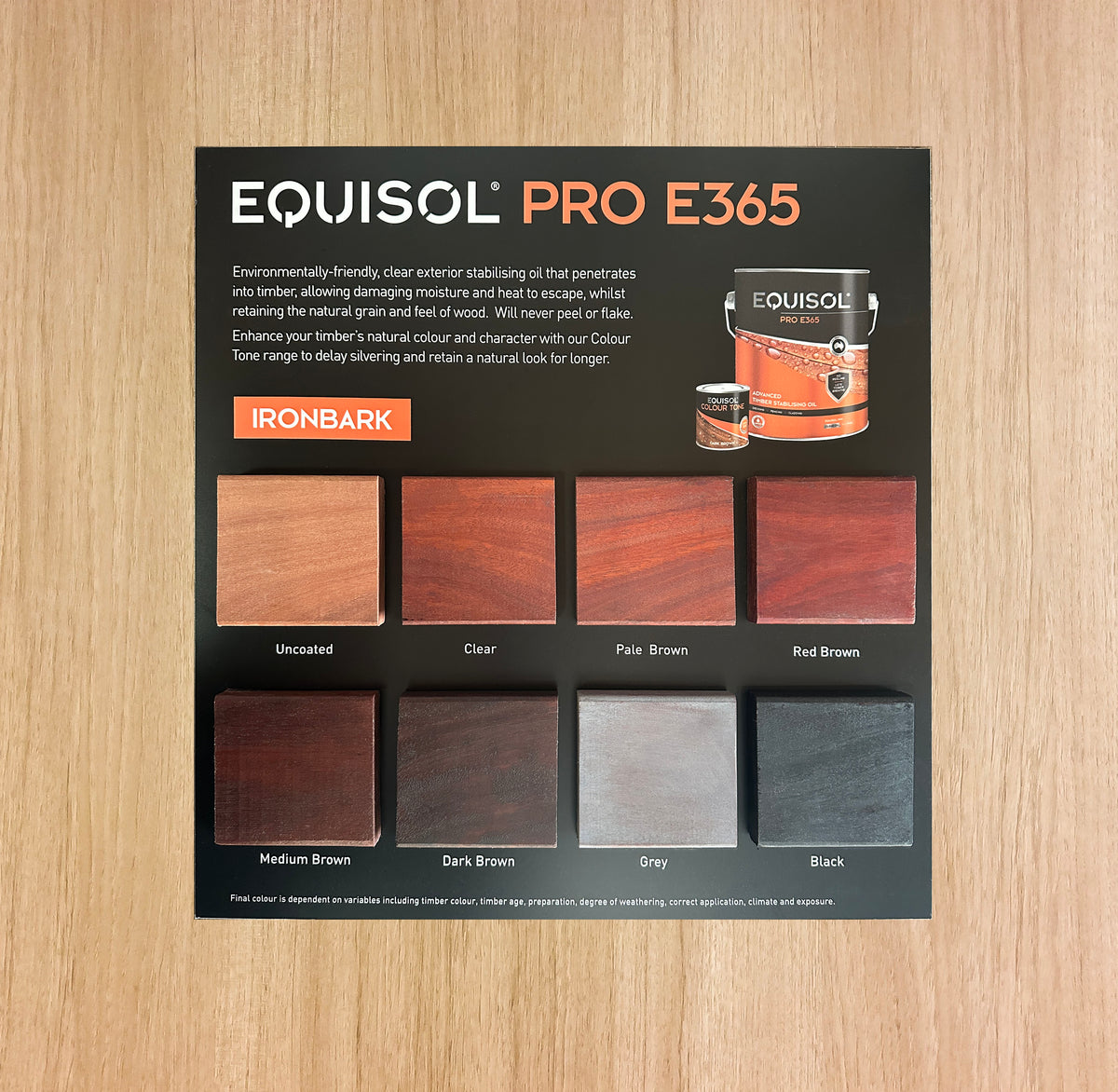 An Equisol Pro E365 ironbark wood finish color card showing various wood swatches from uncoated to black, placed on a light wood grain surface.